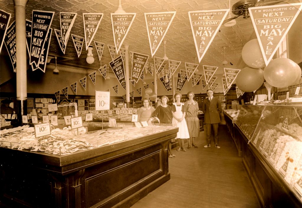A small Woolworth 5 and 10 dominated by a glass candy counter and pennant hanging banners promoting the next Sale event